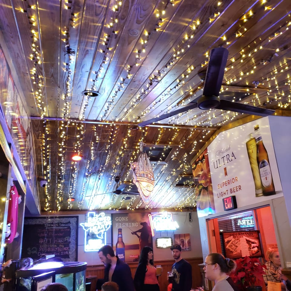 A lively restaurant interior with string lights on a wooden ceiling, people, and neon signs.