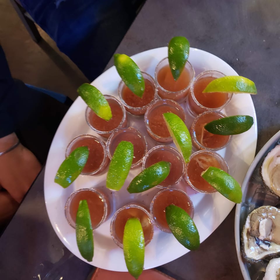 A white platter holds numerous shot glasses filled with a red drink and garnished with lime wedges.